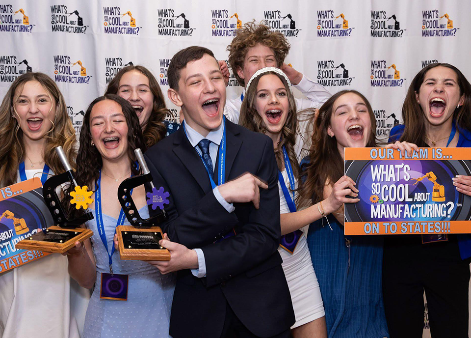 A group of excited pre-teenagers celebrates in front of a "What's So Cool About Manufacturing?" backdrop. They hold plaques and signs, one of which says "Our Team is On to States!!!," while smiling widely with joy.