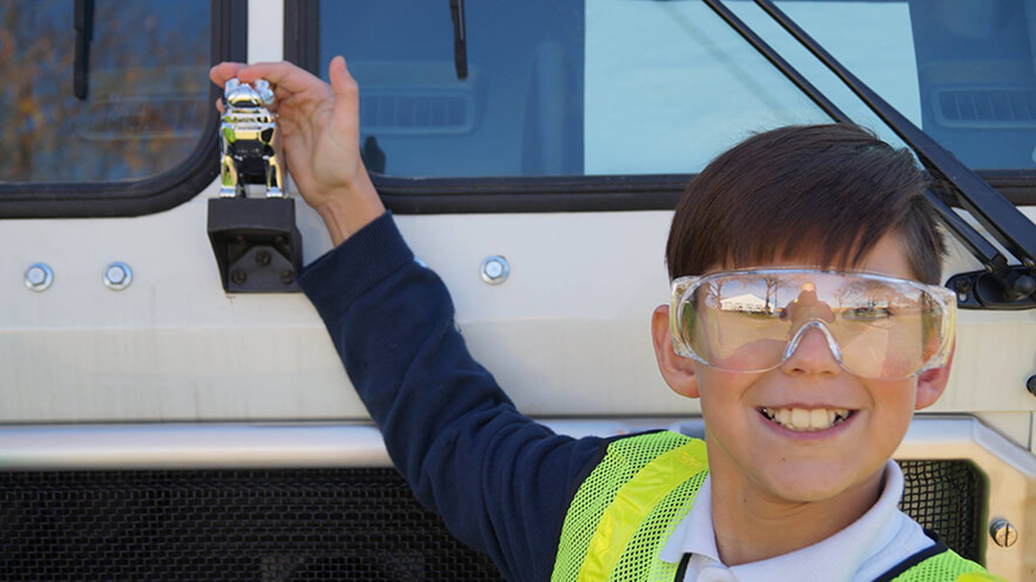 A smiling boy wearing safety goggles and a reflective vest stands next to a Mack Truck.