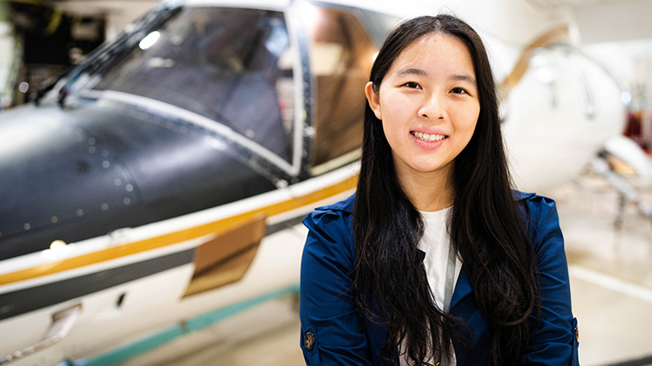A smiling young professional standing confidently in front of a sleek, parked airplane in a hangar, conveying a sense of professionalism.